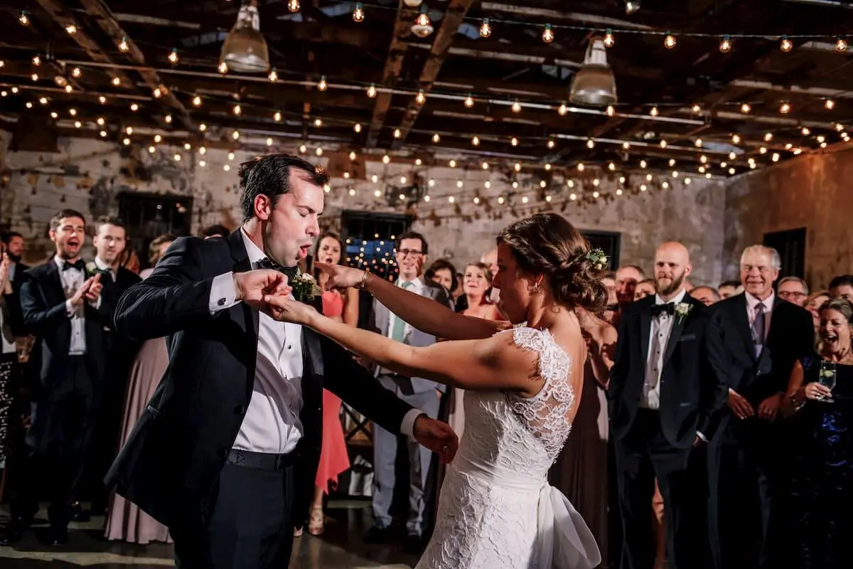 A bride and groom dance under string lights with guests clapping around them