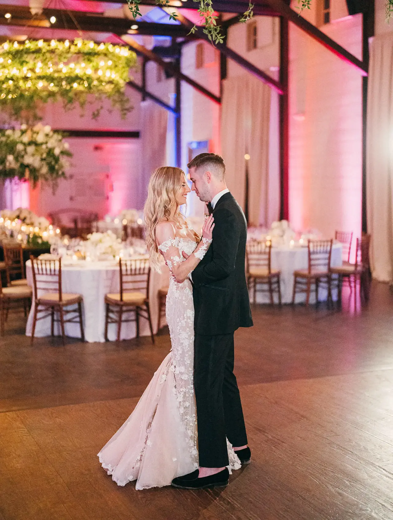 A bride and groom share their first dance in a ballroom lit with pink uplighting